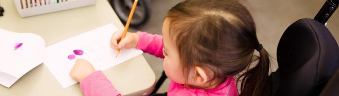 Little girl in a wheelchair at a table writing on a paper with pencil